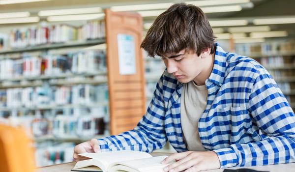 En tenåringsgutt sitter ved et bord på et bibliotek og leser. Bilde.