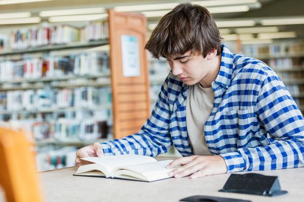 En tenåringsgutt sitter ved et bord på et bibliotek og leser. Bilde.