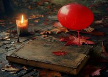 Una novela de terror de tapa dura de los años ochenta sobre una mesa, junto a un globo rojo y hojas de otoño, iluminada por velas
