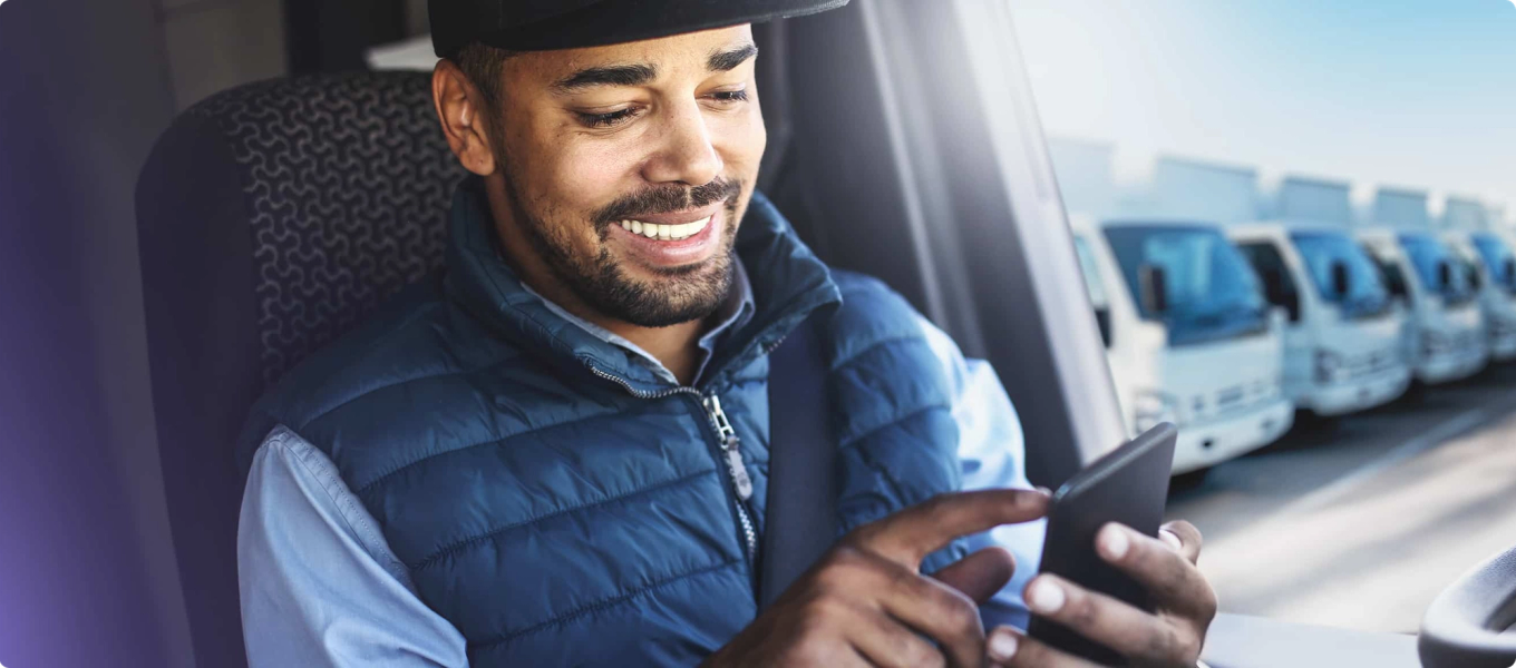 Man in a truck scrolling on his phone with a fleet of trucks visible in the background.