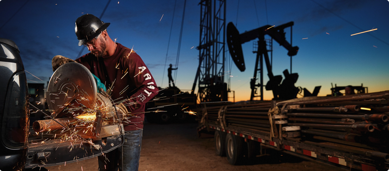 Worker using a circular saw to cut metal at an oil site, with sparks flying at dusk.