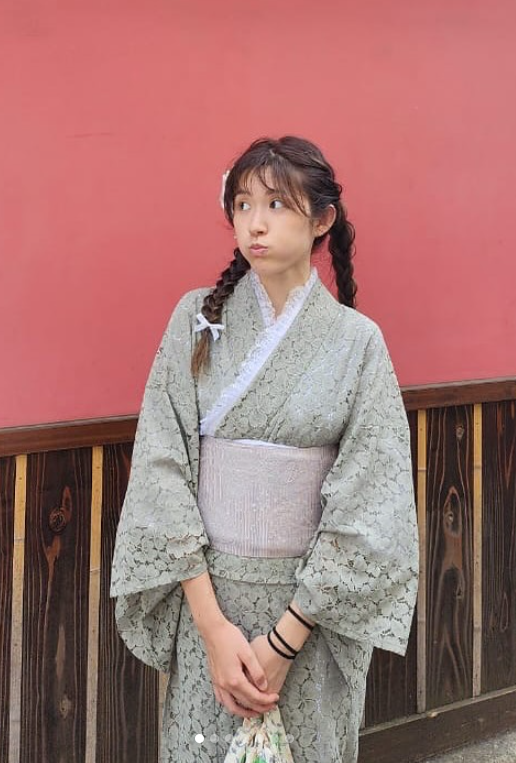 Yuriko Goto wearing a sage-green lace kimono, standing in front of a red wall and wooden lattice fence
