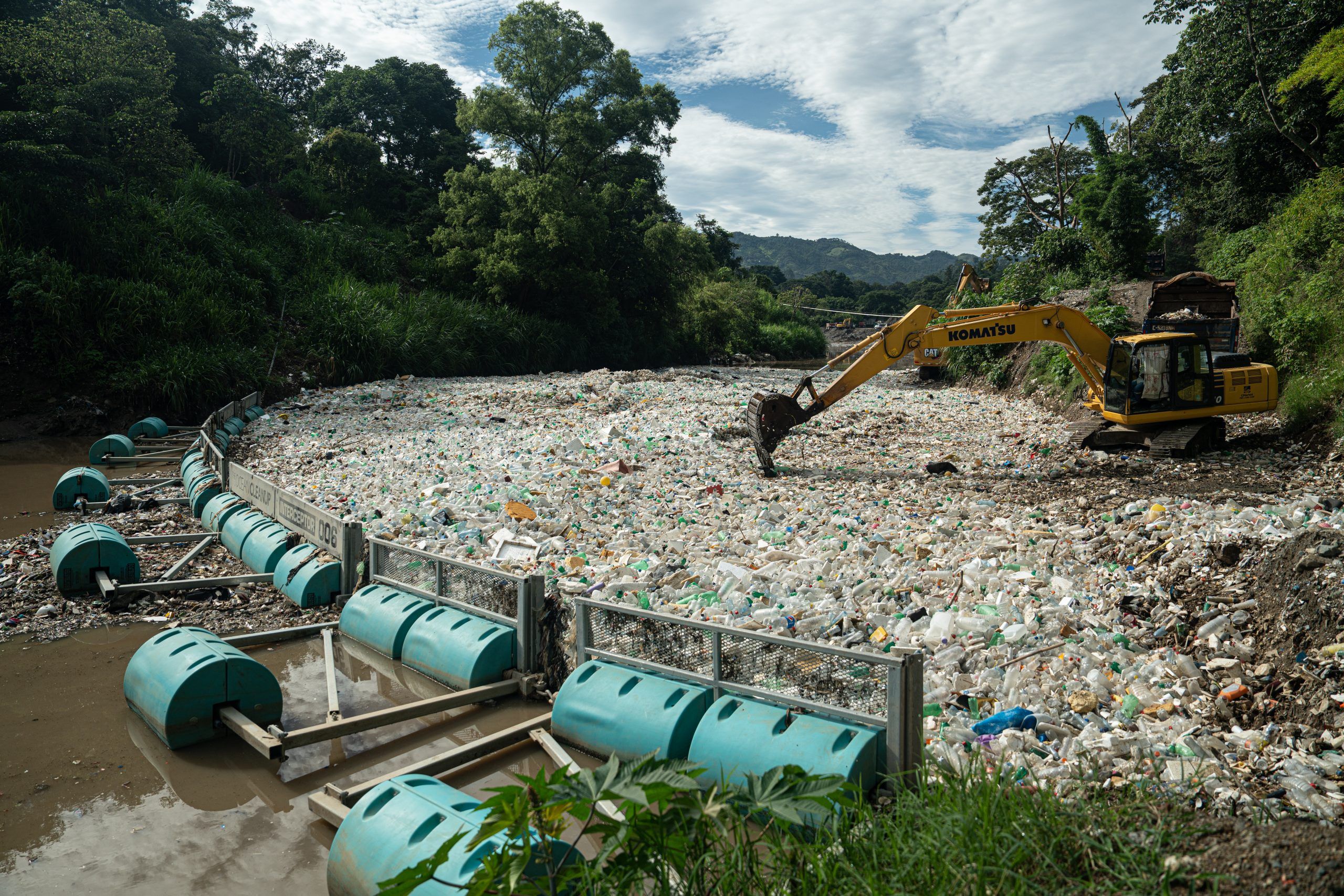 Excavator clearing plastic waste from a river blocked by a floating barrier.