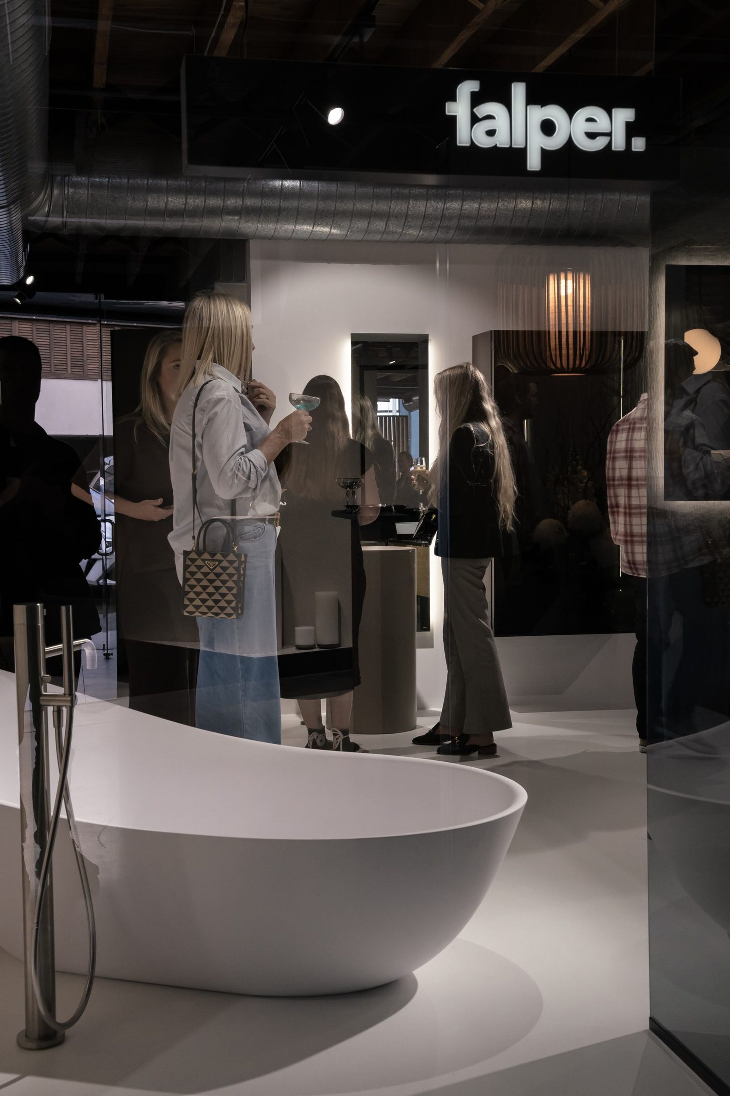 Evening showroom event seen through a glass partition: guests in conversation, one raising a cocktail coupe, behind a sculptural white freestanding bath with a floor-mounted mixer and handshower, beneath an illuminated 'falper.' sign