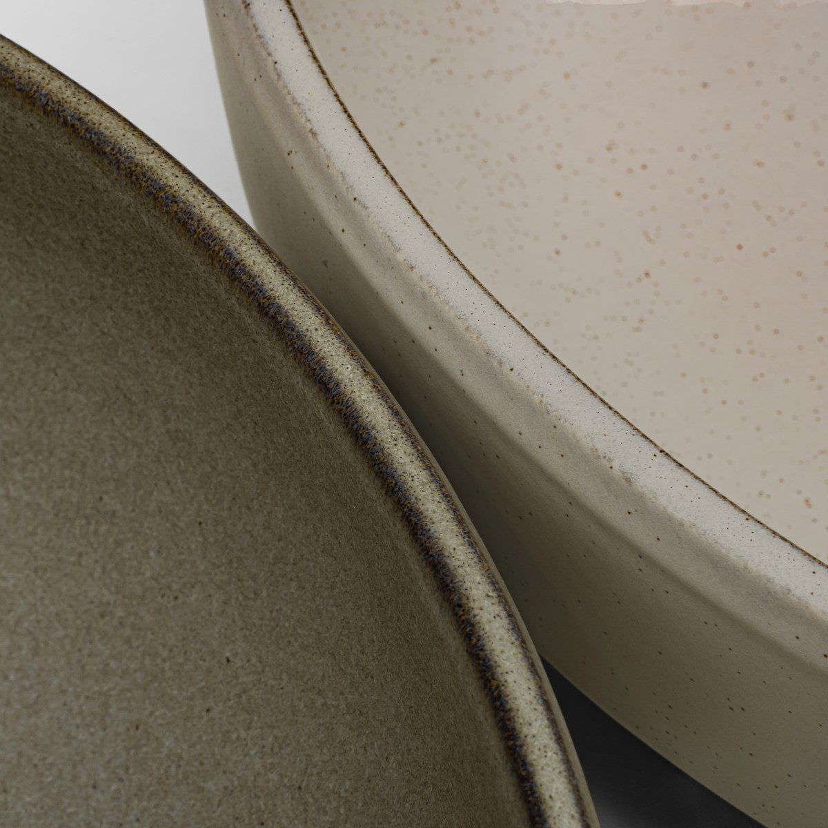 Macro close-up of two overlapping ceramic vessels, a matte olive-green bowl resting against a speckled oatmeal one, with dark reactive glaze breaking along each rim