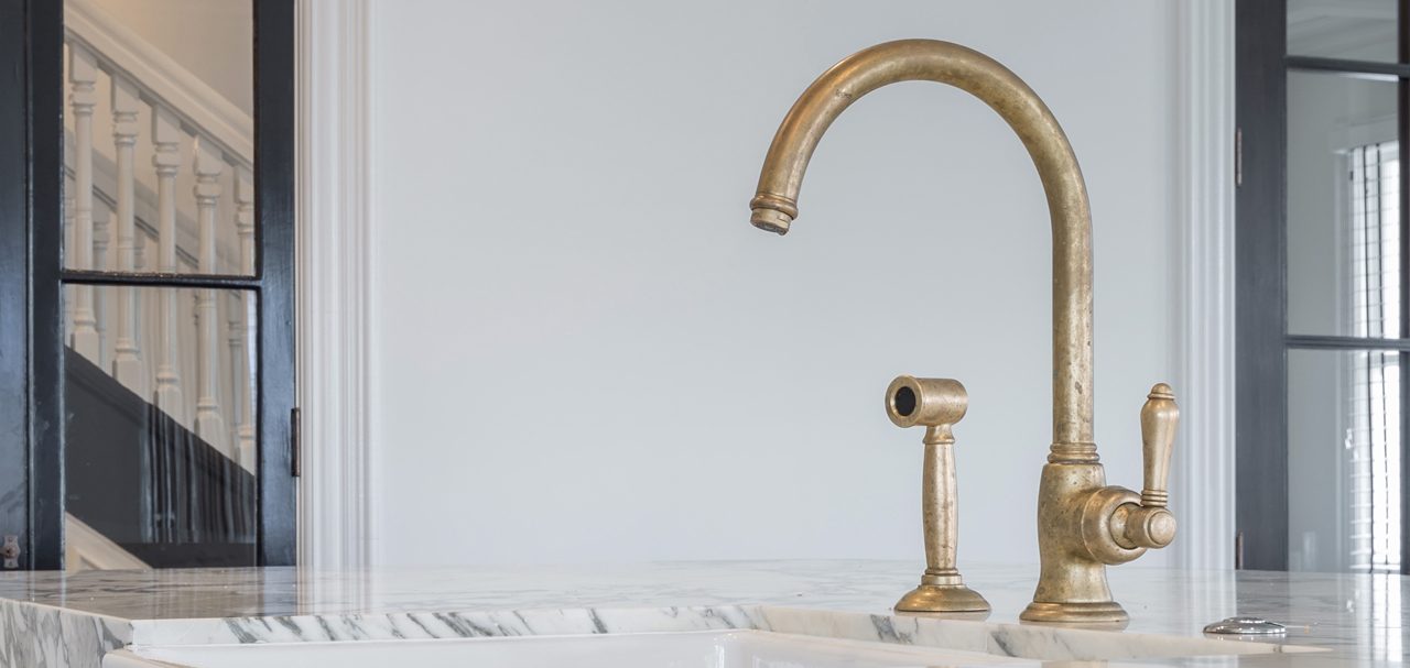 Aged brass kitchen mixer and spray on a marble benchtop above a white butler sink