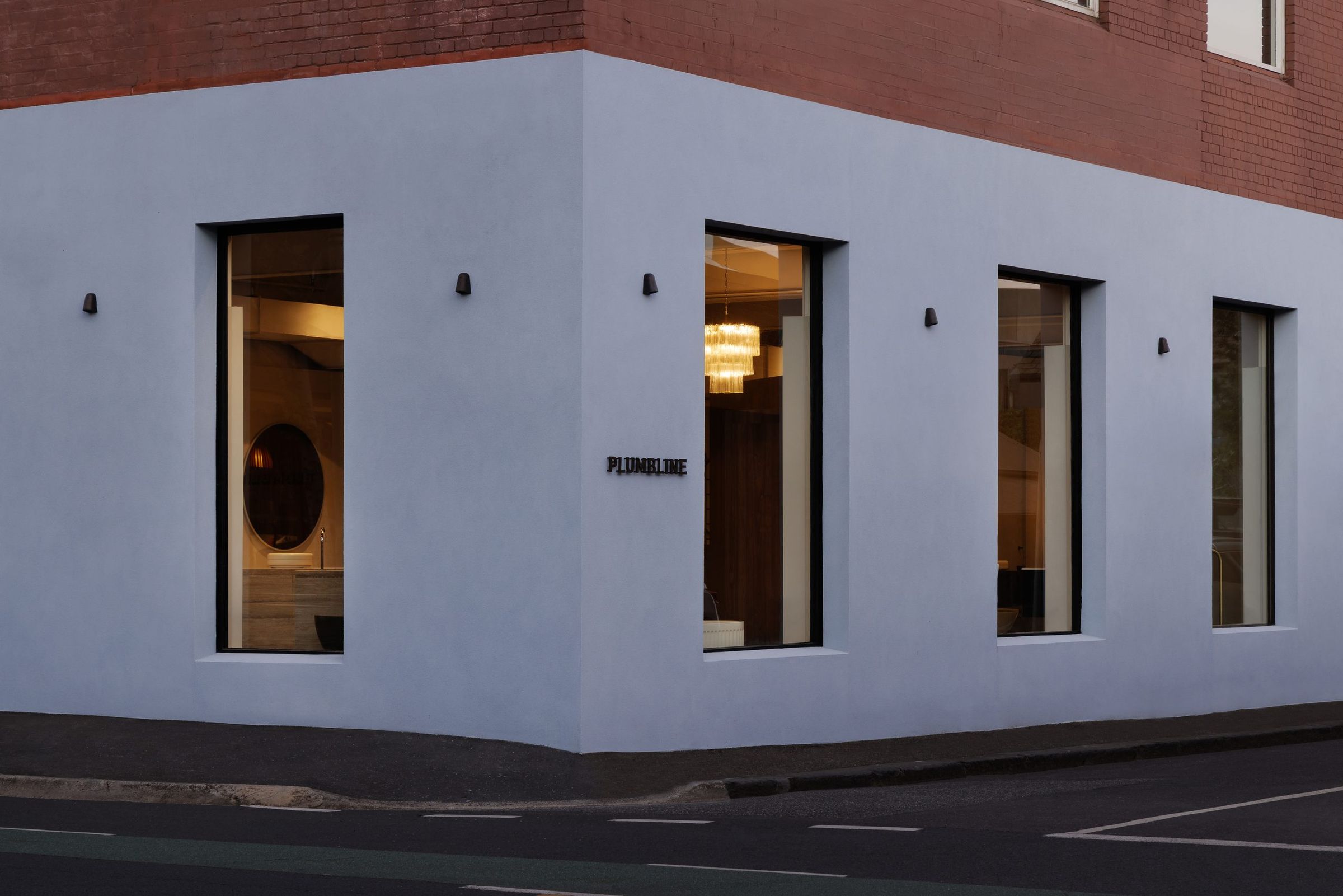 Plumbline's showroom on a street corner at dusk — a pale lilac rendered facade wrapping the corner beneath red brick, tall black-framed windows glowing warm from within, where a fluted glass chandelier, a round mirror and a timber vanity with a dark basin are visible, and the PLUMBLINE nameplate mounted on the corner wall above an empty road.