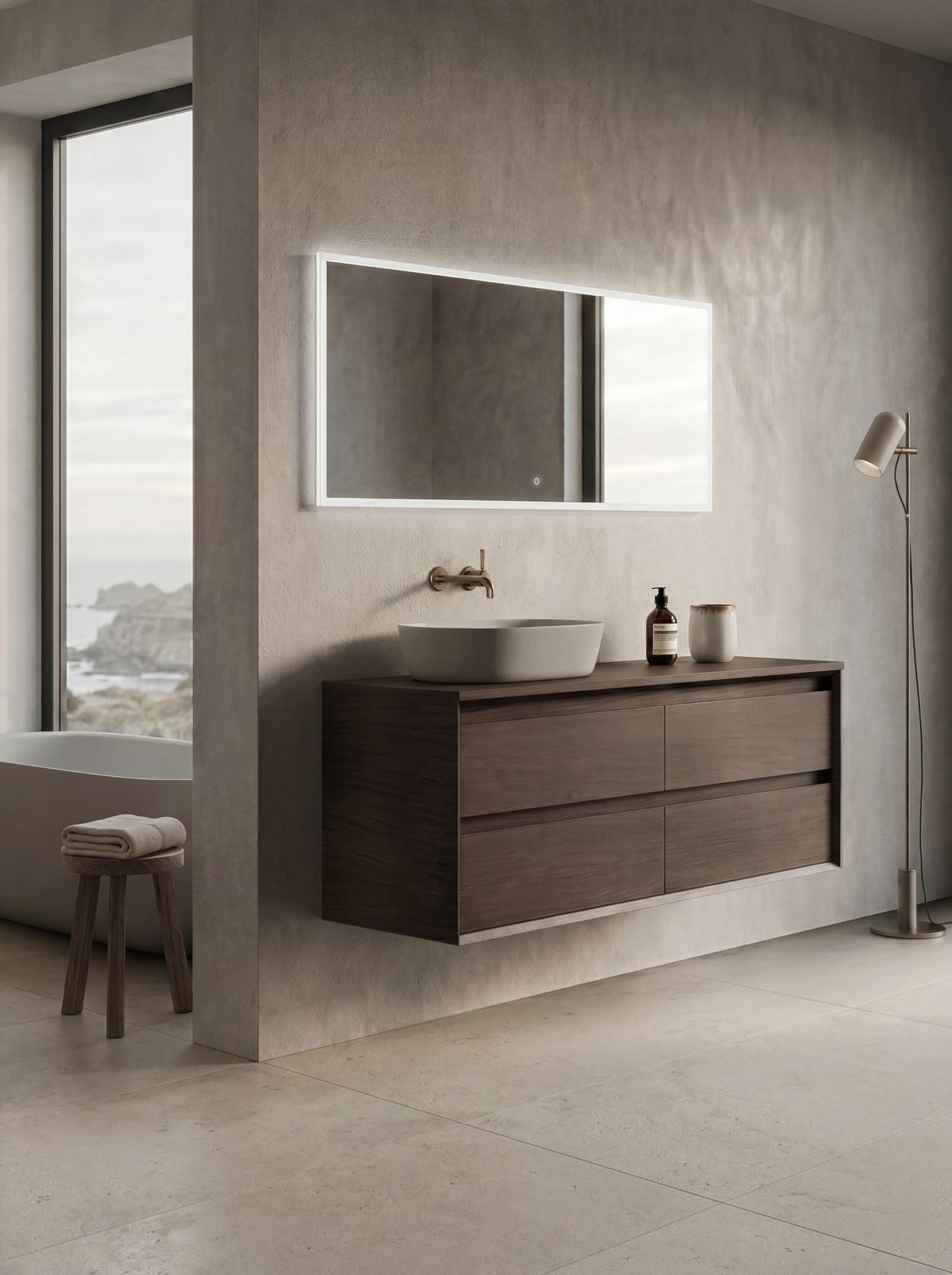 Wall-hung dark timber vanity with a round stone basin and bronze wall-mounted tapware beneath a backlit LED mirror, in a coastal bathroom overlooking a rocky shoreline