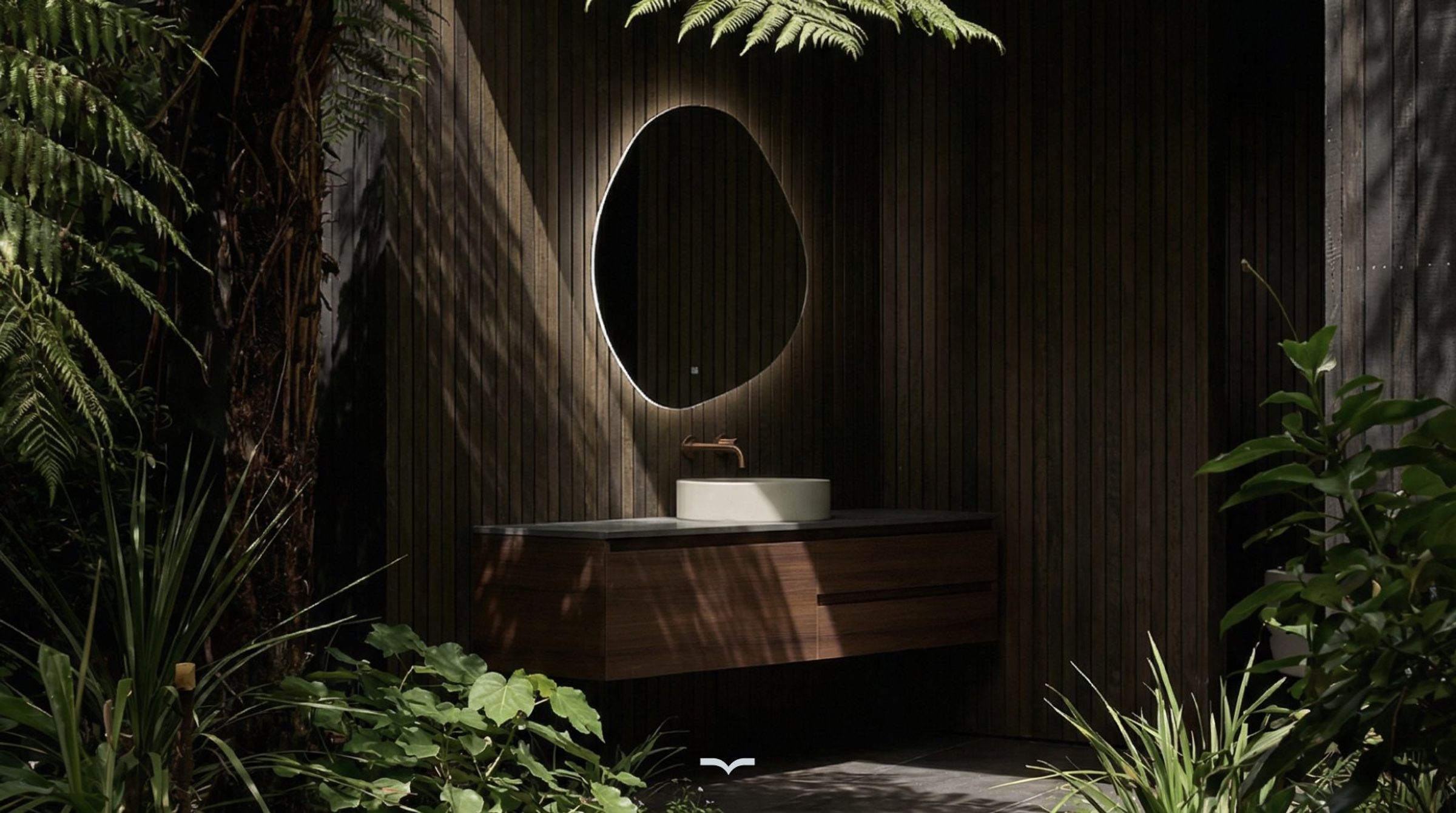 A dark timber-battened outdoor bathroom framed by native ferns and broadleaf foliage, with dappled sunlight falling across a walnut floating vanity, stone benchtop, pale round vessel basin and wall-mounted bronze tapware beneath a rim-lit pebble-shaped mirror.