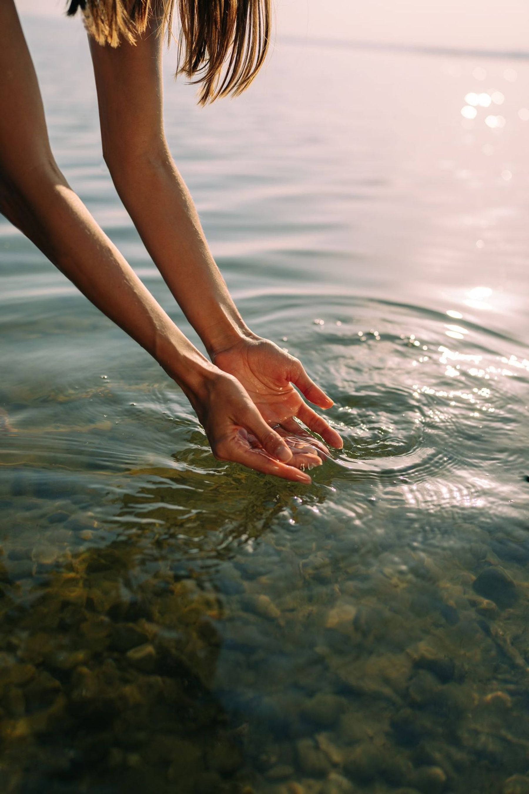 Cupped hands dipping into clear shallow water at golden hour, ripples spreading over a sunlit pebble bed