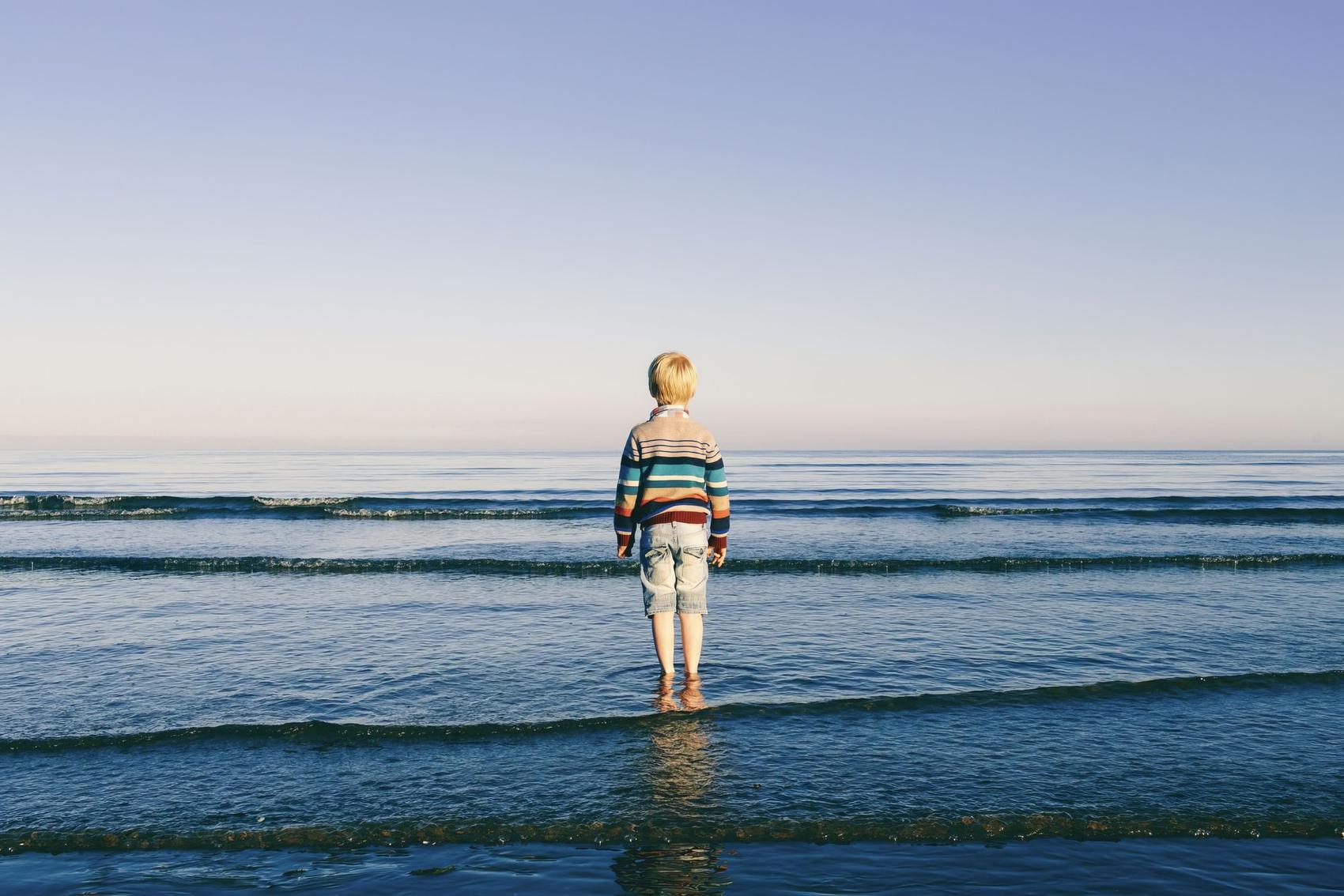 A young child in a colourful striped jumper and denim shorts stands ankle-deep in calm shallow sea, back to camera, looking out past low breaking waves to a pale horizon
