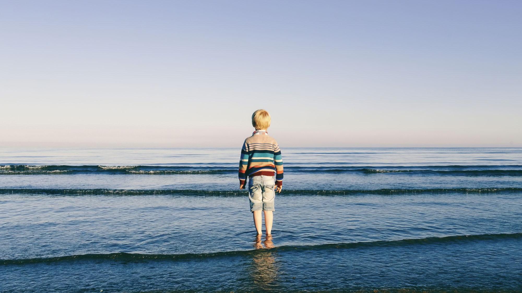 A young blond child stands alone and ankle-deep in calm shallow sea, seen from behind in a multicoloured striped knit jumper and pale denim shorts, facing a flat open horizon under a soft lilac dusk sky