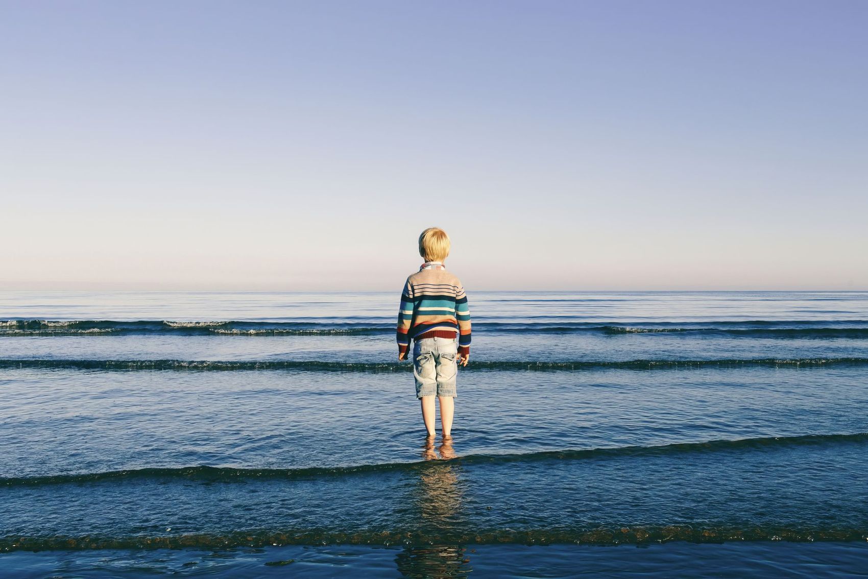 A young child in a colourful striped jumper and denim shorts stands ankle-deep in calm shallow sea, back to camera, looking out past low breaking waves to a pale horizon
