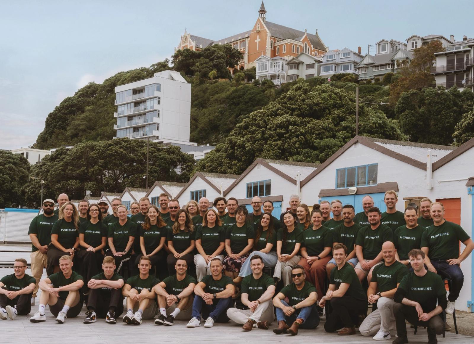 The Plumbline team, around fifty people in dark green branded T-shirts, gathered in rows on the Wellington waterfront in front of the white Clyde Quay boat sheds, with a bush-covered hillside of timber villas and a brick church above them