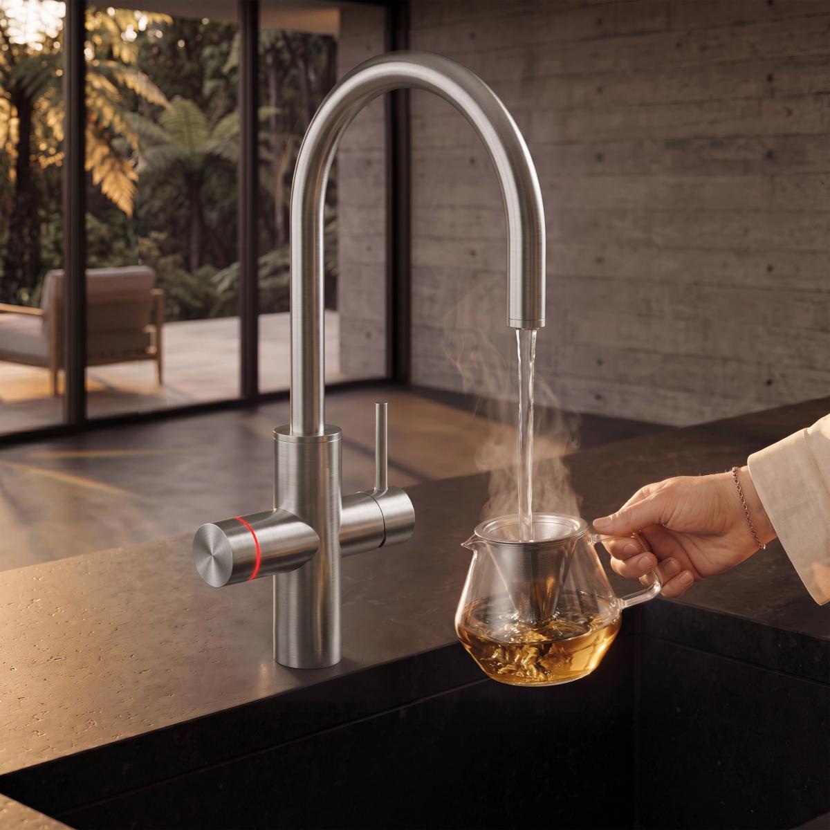 Brushed brass twin-handle boiling-water kitchen mixer pouring a steaming stream into a matte black ceramic teapot held by a woman's hand, on a dark stone benchtop against a board-formed concrete wall, with native tree ferns beyond the glazing