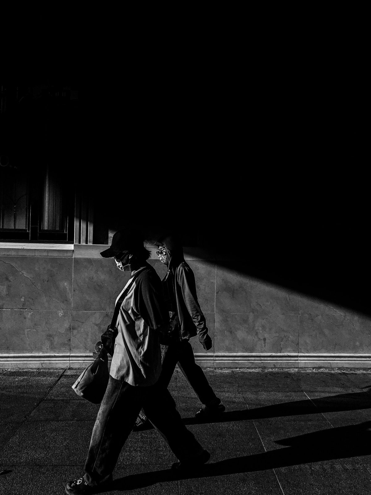 Dos figuras caminando, recortadas por una franja de luz sobre un muro en sombra