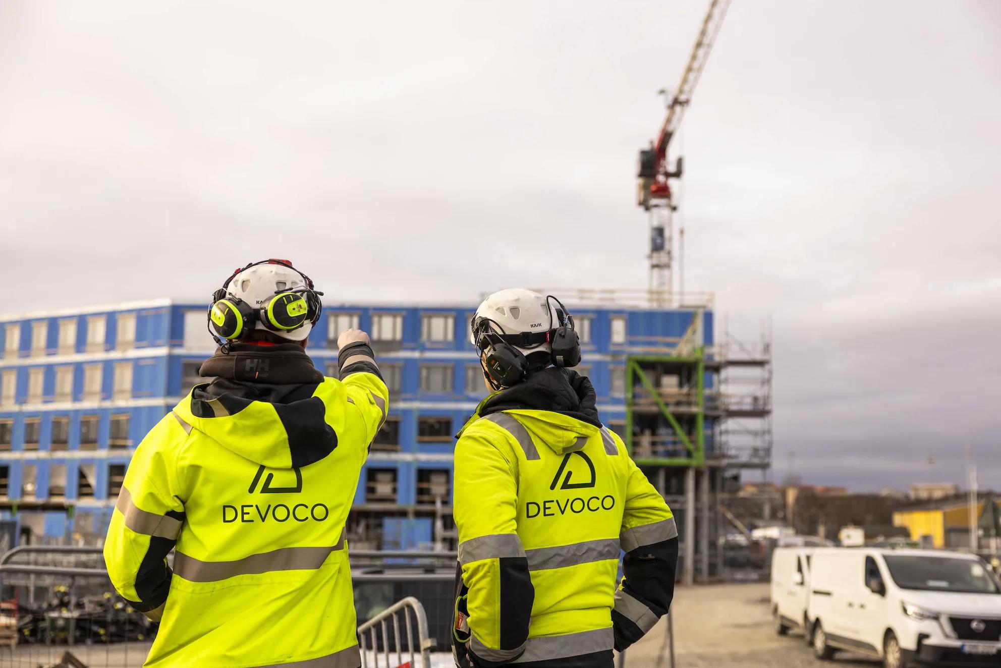 Two people in Devoco high-visibility jackets on a building site, pointing at a building under construction with a tower crane behind it
