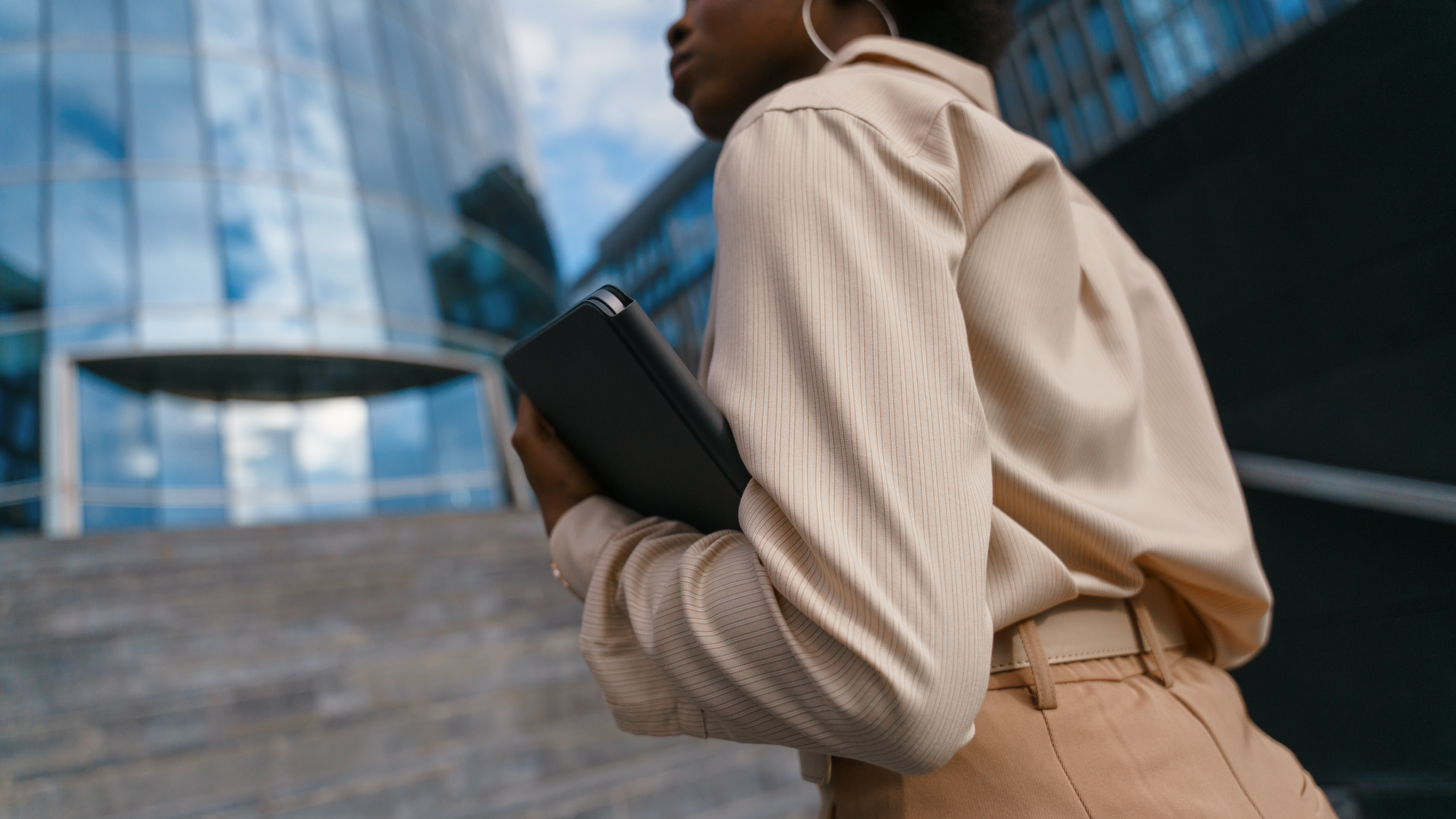 A person holds a tablet while walking up concrete stairs towards a modern glass building.