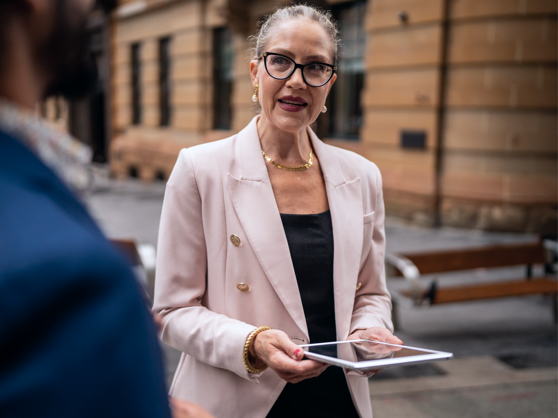Woman in a pink blazer and glasses holds a tablet while talking with a colleague outdoors