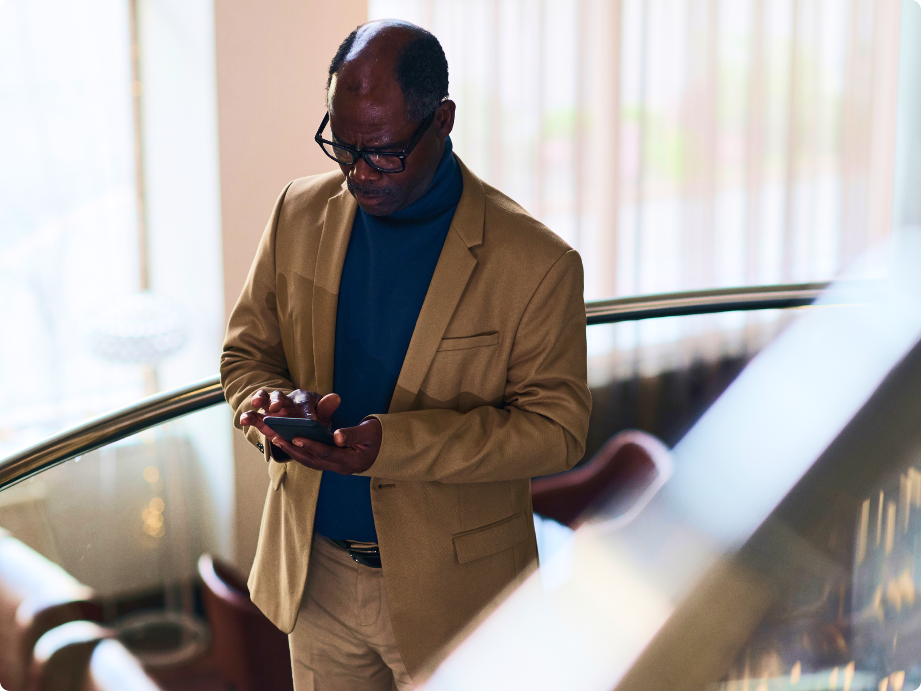 An older Black man in a tan blazer and glasses stands on an escalator while using his smartphone.