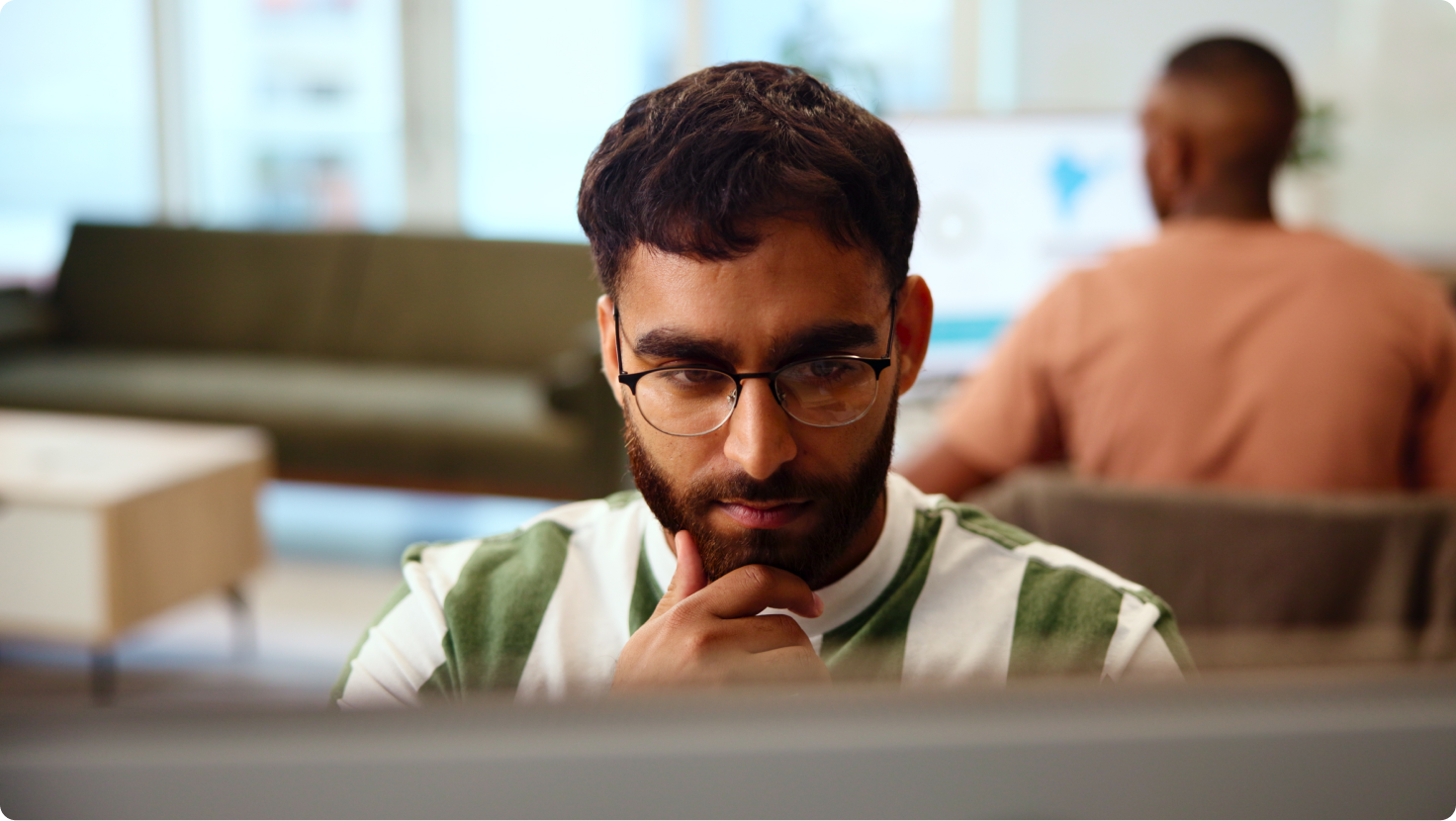 A thoughtful man with glasses and a beard looks intently at a screen, hand on chin, in an office.