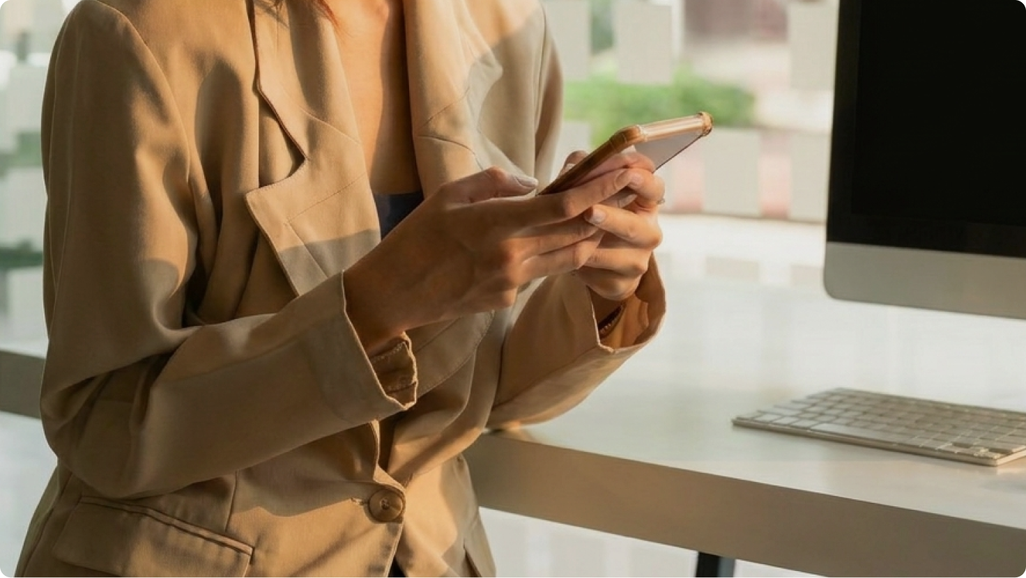 Close-up of a person in a beige blazer using a smartphone at an office desk.