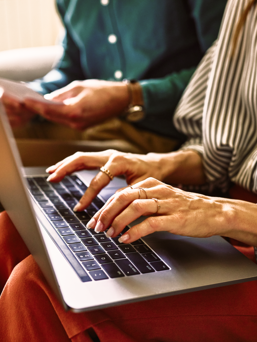 Close-up of hands typing on a laptop keyboard, with another person holding documents in the background.