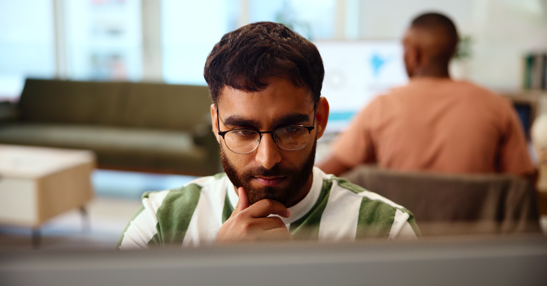 Man with a beard and glasses looks intently at a screen, hand on chin.