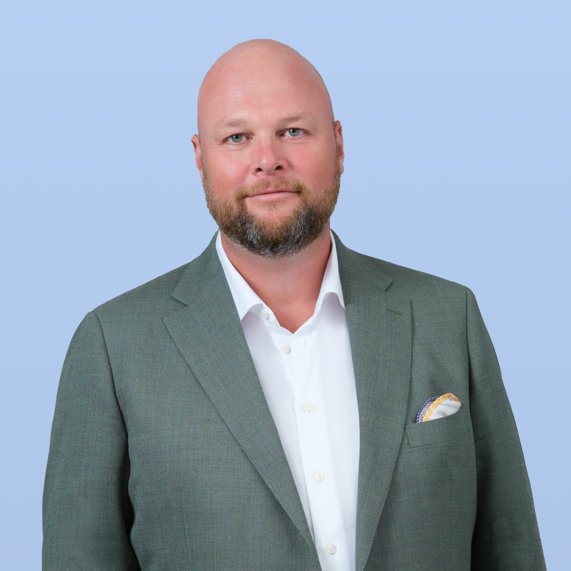 A bald man with a beard wears a green suit jacket, white shirt, and patterned pocket square, looking forward against a light blue background.