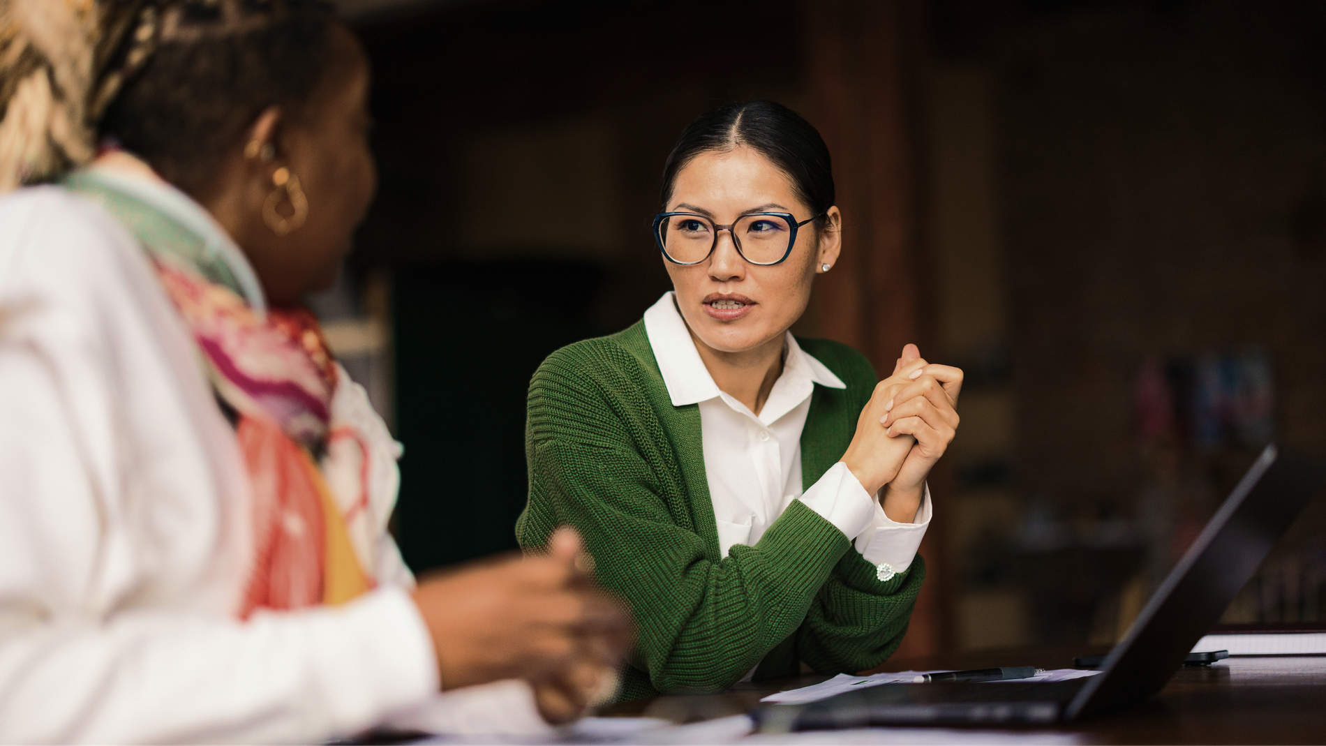 An Asian woman in glasses and a green cardigan listens attentively to another person across a table with a laptop.