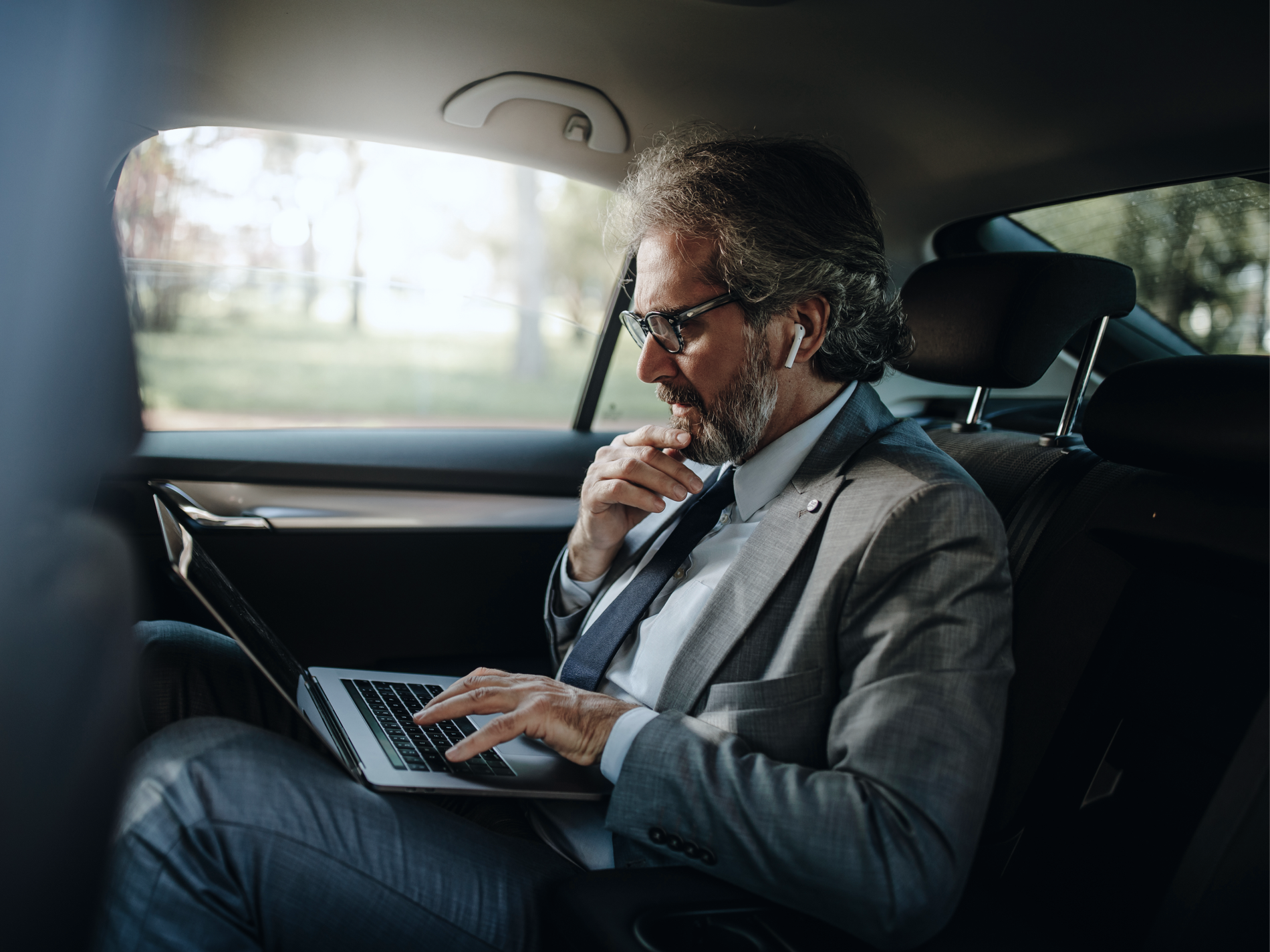 Businessman in a suit typing on a laptop in the back of a car.