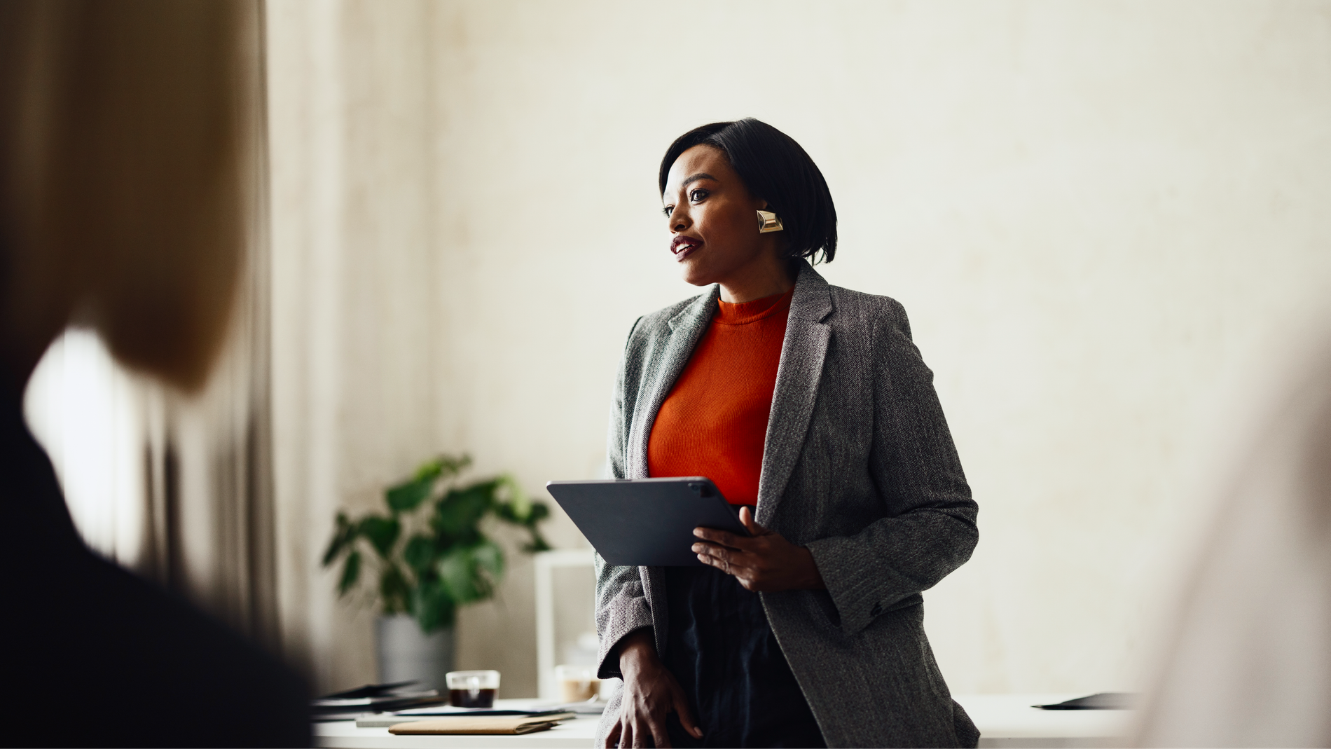 A professional in a grey blazer holding a tablet while speaking to colleagues in a bright office.