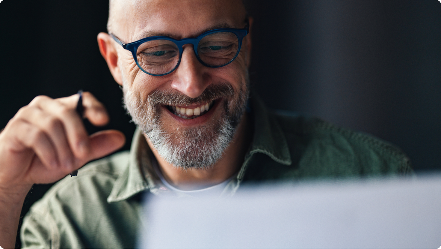 Smiling man with a beard and blue glasses looks down, holding a pen.