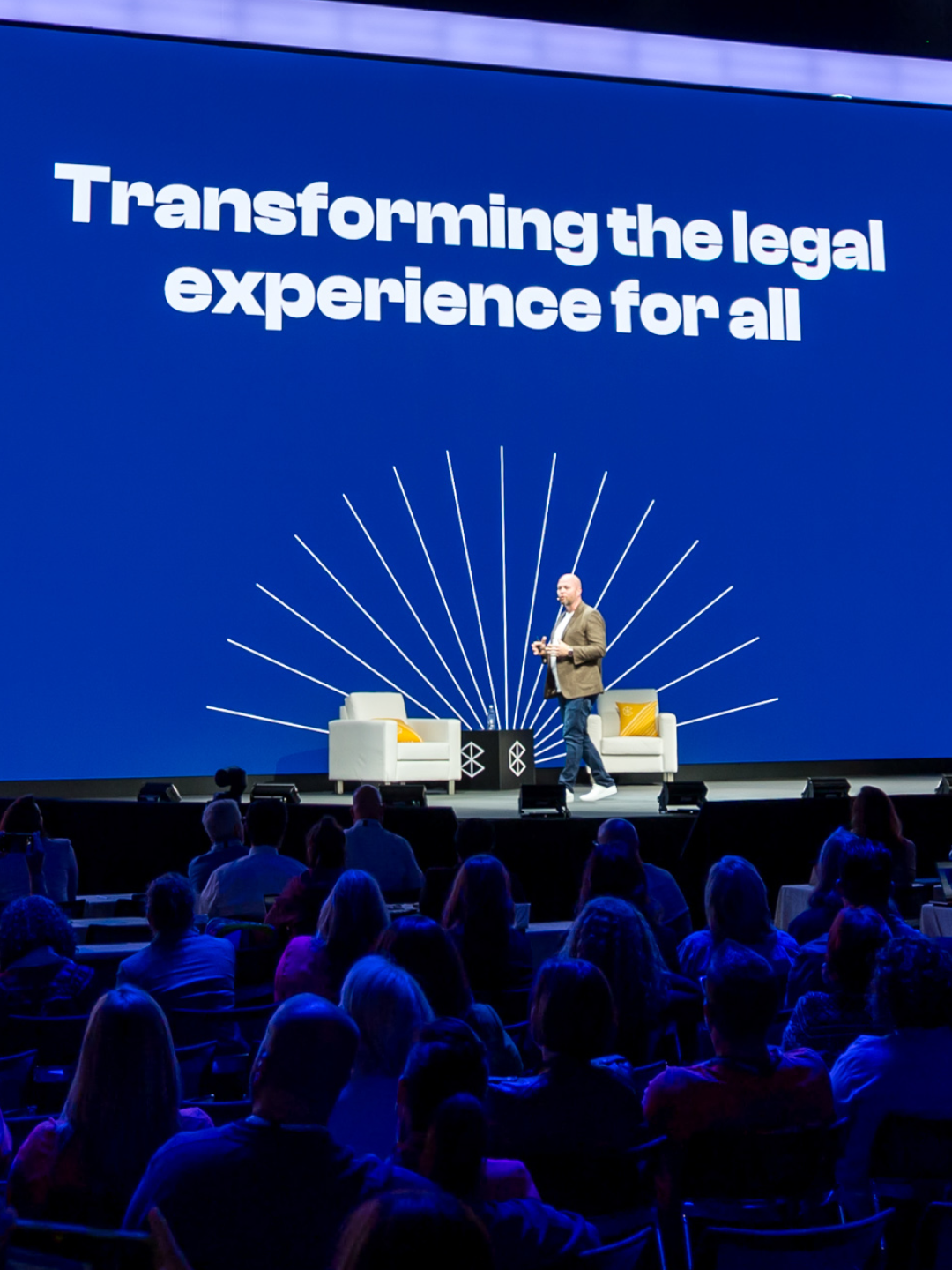 A man presents on stage in front of a blue screen that reads "Transforming the legal experience for all" to an audience.