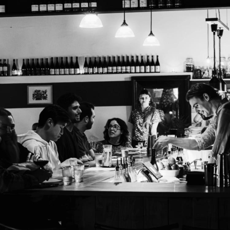Black and white photo of a bartender serving drinks to customers seated at a bar, showed for the Clio event at Brisbane.