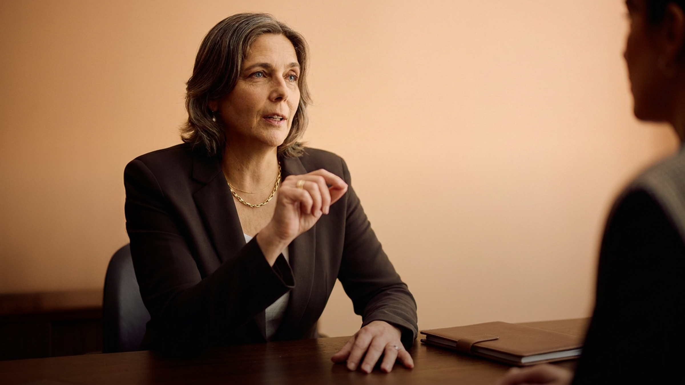 A businesswoman in a suit gestures while speaking to someone across a desk.