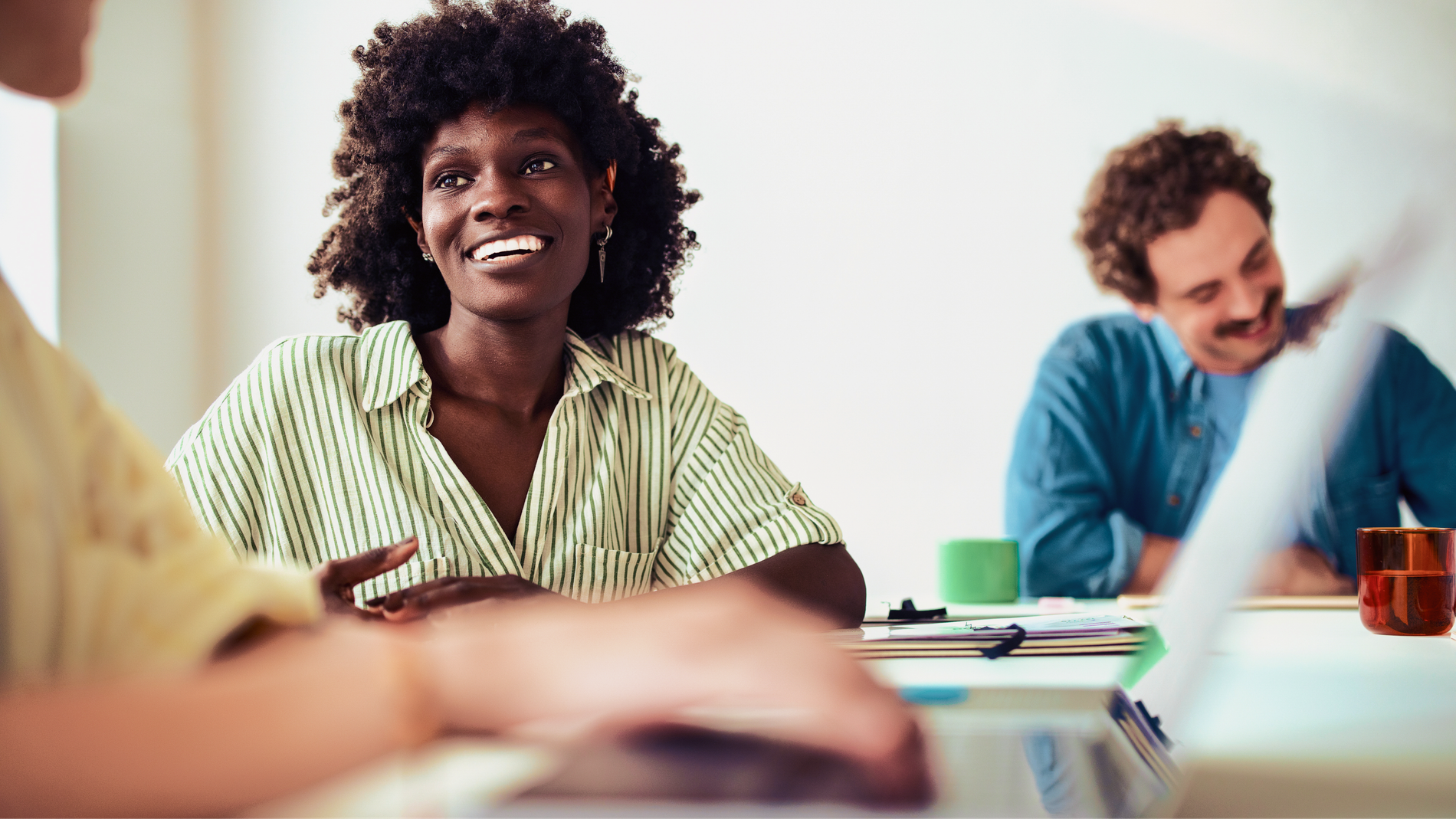 Smiling woman in a striped shirt talking with colleagues across a bright meeting table.
