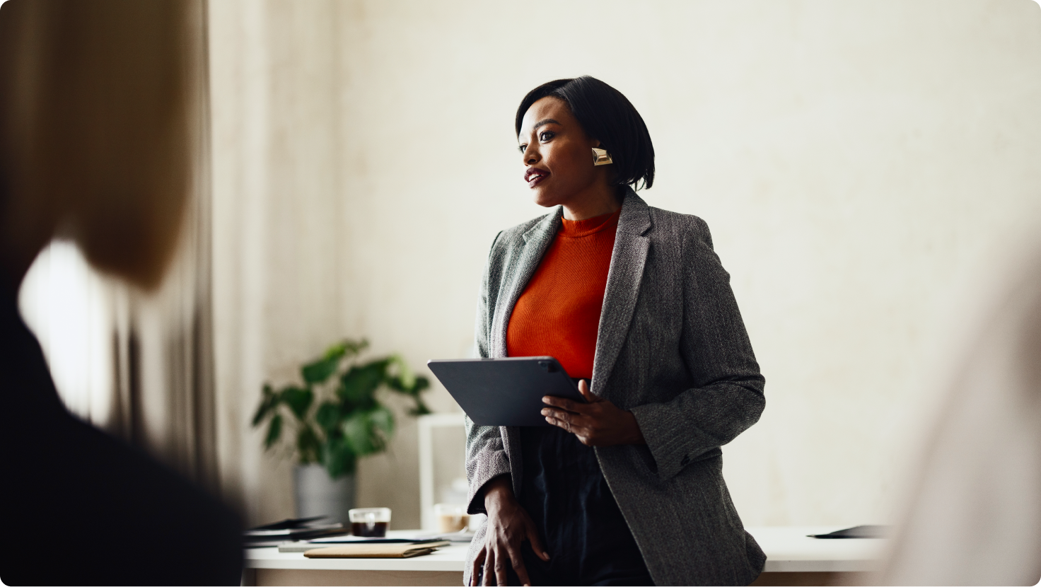 A Black woman in a blazer holds a tablet and speaks to an unseen audience.