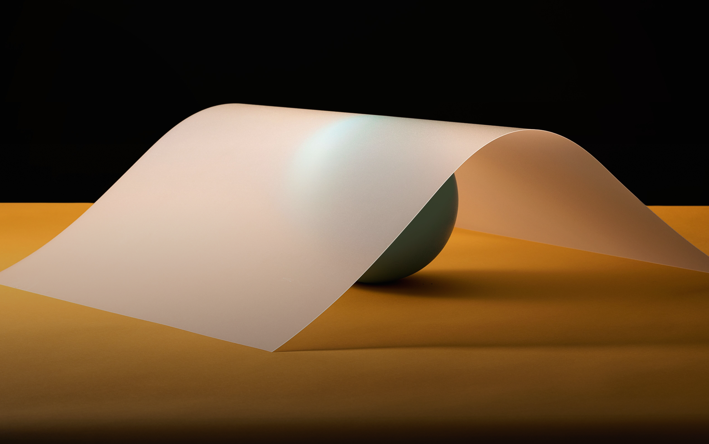A translucent sheet draped over a green sphere on a yellow surface against a black background.
