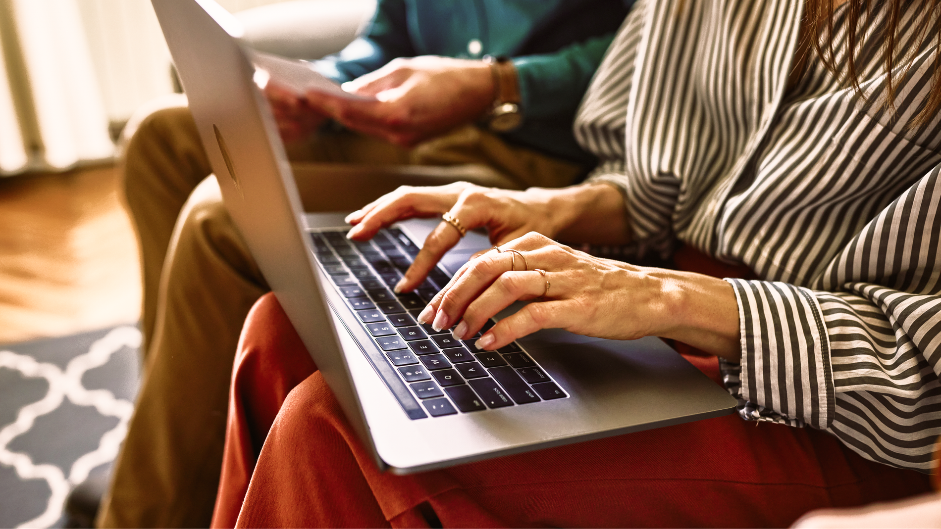 Two people on a sofa, one typing on a laptop while the other reviews a printed document.