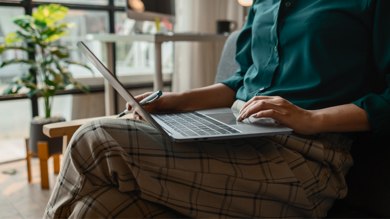 Person in a green shirt and plaid pants using a laptop, holding a pen.