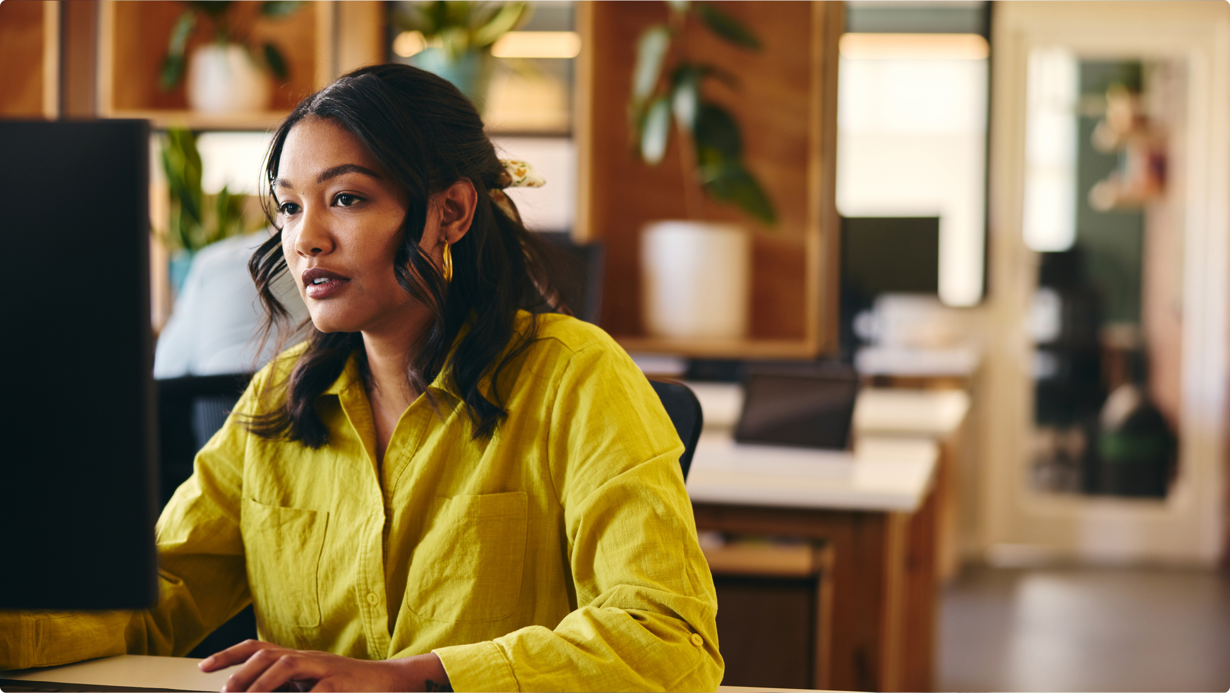 A young woman in a yellow shirt works intently at a computer in an office.