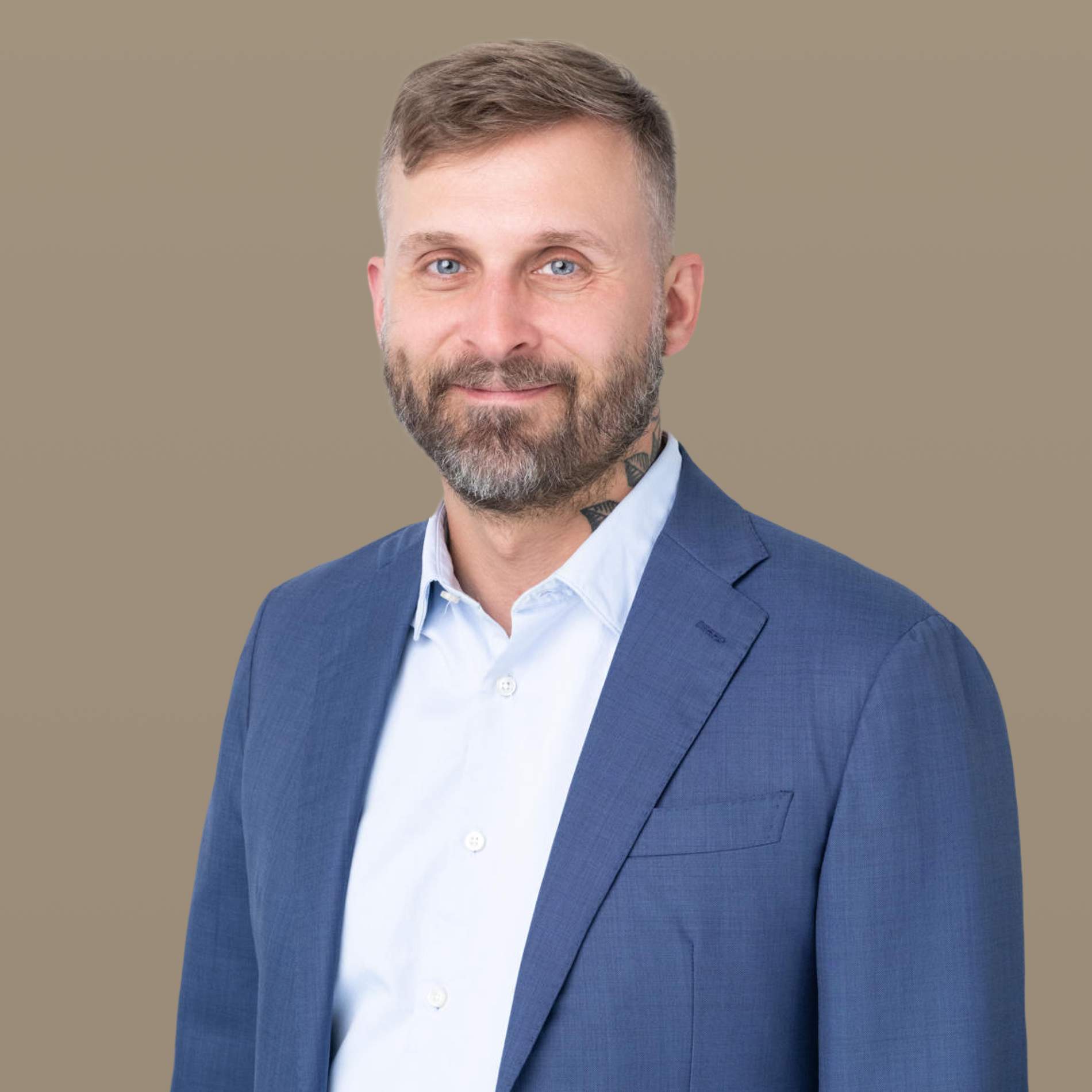 Headshot of a smiling man with a beard and neck tattoo, wearing a blue suit jacket and light blue shirt, against an olive green background.
