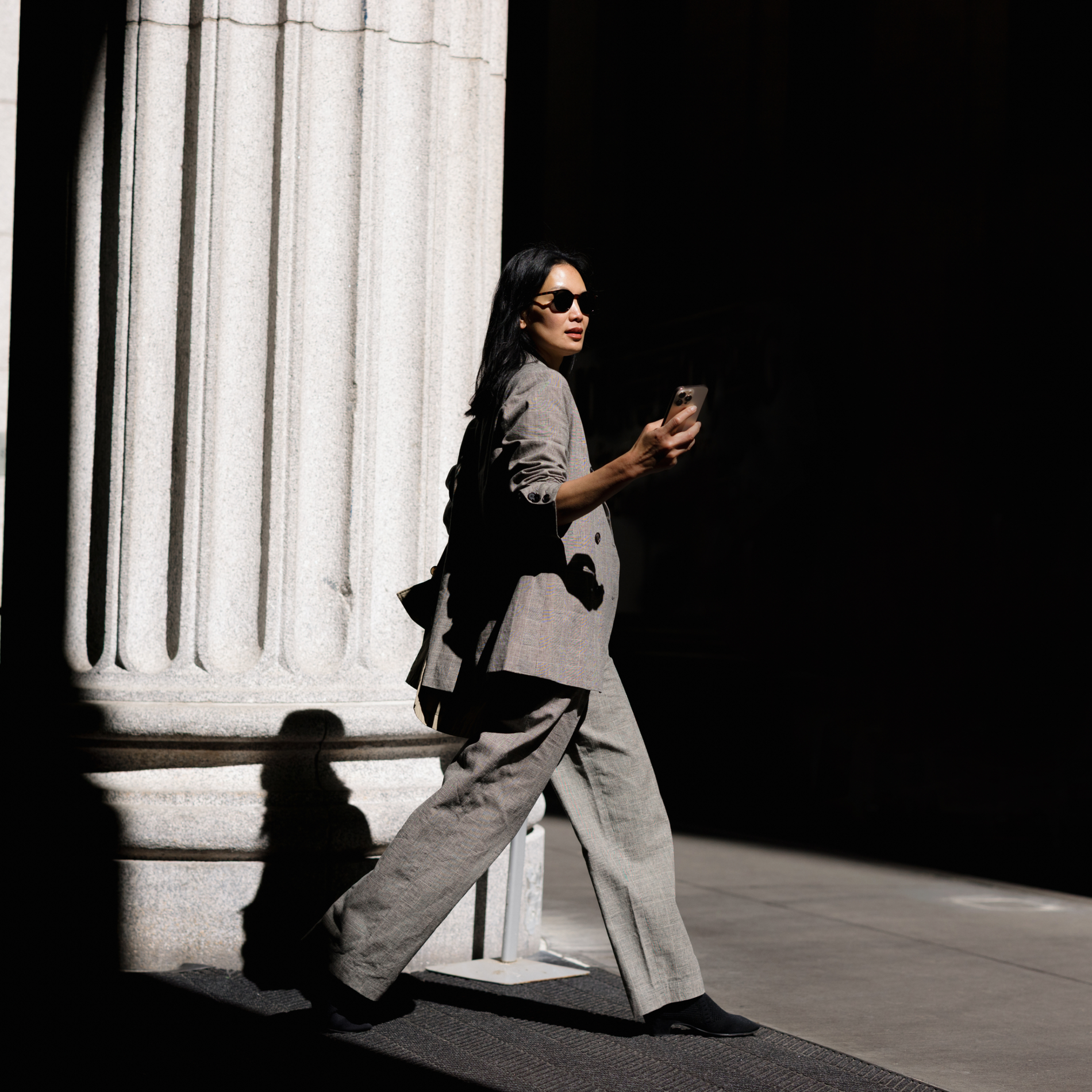 A woman in a gray pantsuit and sunglasses walks past a stone column, holding a phone, with dramatic lighting.