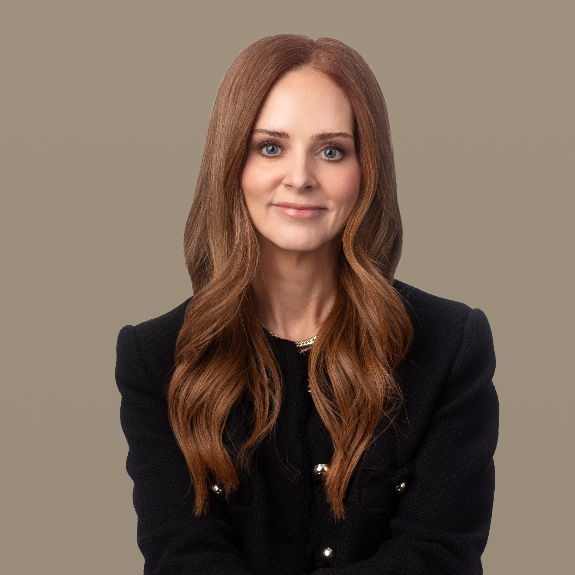 A smiling woman with long reddish-brown hair, wearing a white blazer, stands with arms crossed against a purple background.