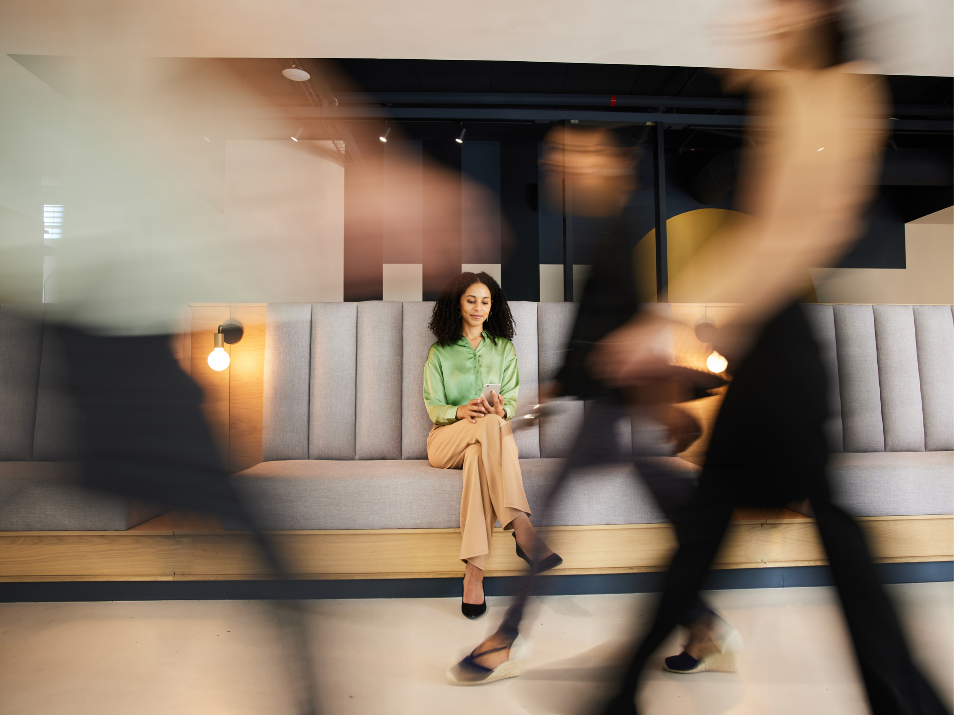 Woman in a mint blouse sits calmly on a lobby bench checking her phone as people hurry past