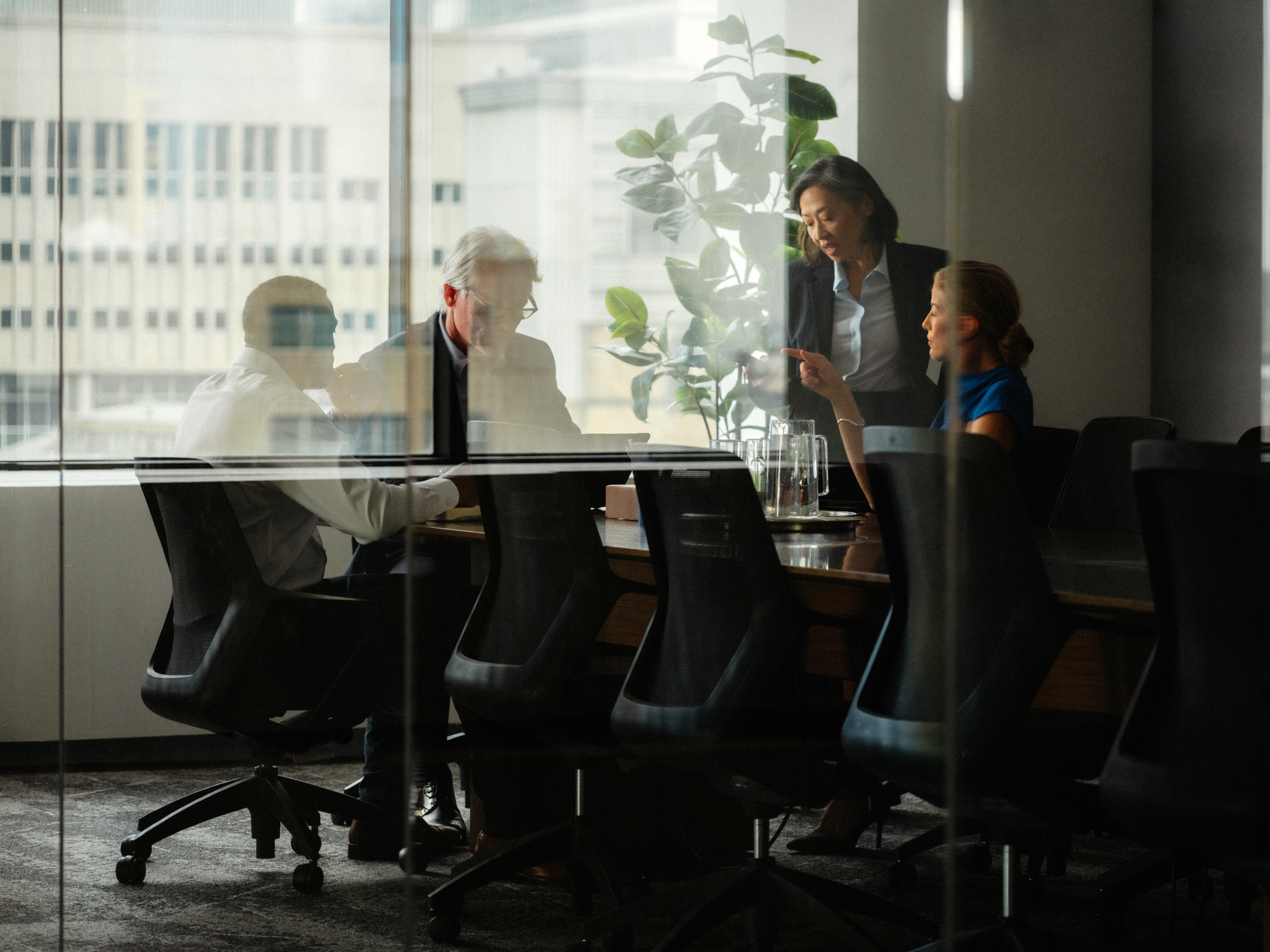 A business meeting with four people in a modern office, viewed through reflective glass, showing city buildings in the background.