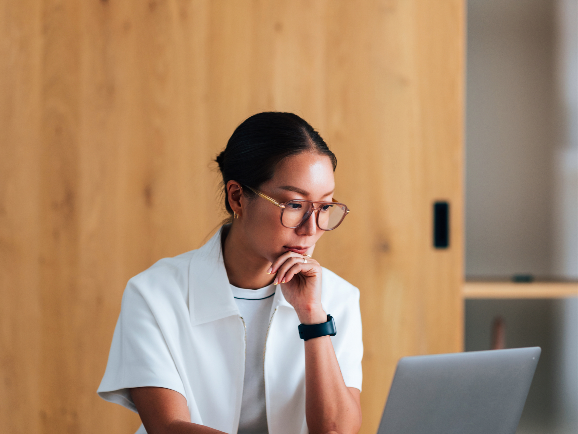Woman in glasses with hand on chin looking thoughtfully at a laptop.