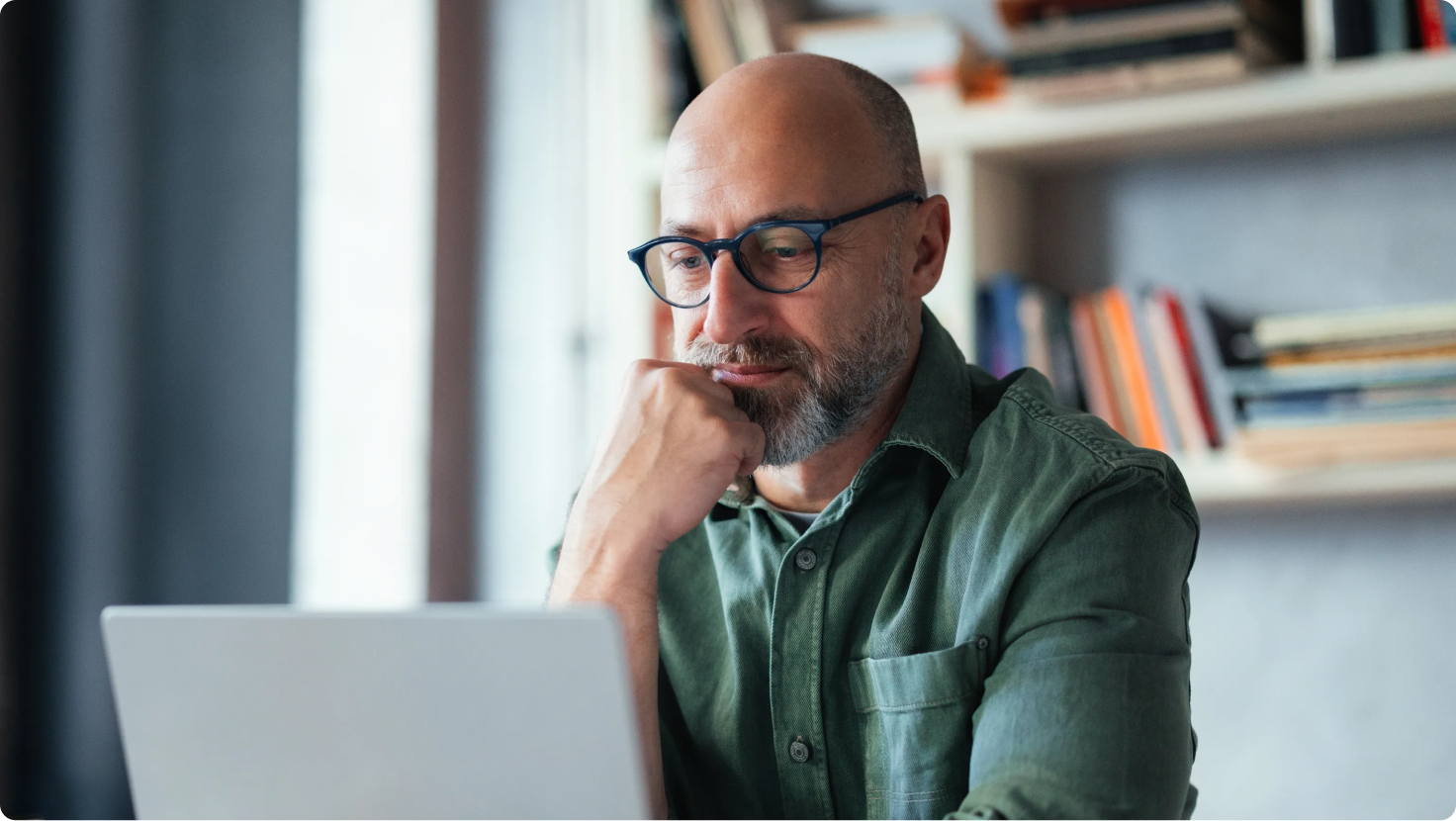 Bald man with beard and glasses looking thoughtfully at a laptop, hand on chin.