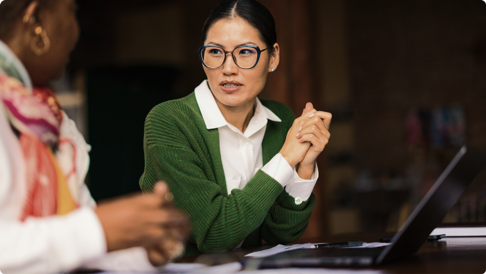 An Asian woman in glasses and a green cardigan sits with clasped hands, engaging in conversation with another person across a table with a laptop.